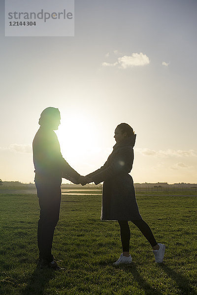 Zärtliches junges Paar  das bei Sonnenuntergang Hand in Hand auf einer Wiese steht.