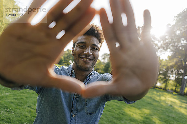 Porträt eines lächelnden jungen Mannes  der im Park einen Fingerrahmen macht.