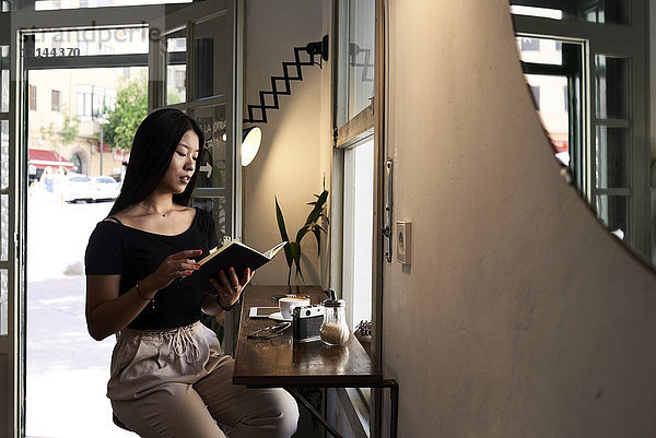 Hübsche asiatische Brünette beim Lesen eines Buches in einem Café am Fenster.