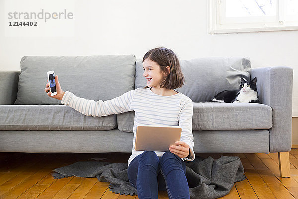 Fröhliches Mädchen mit Tablett auf dem Boden des Wohnzimmers sitzend  das sich mit dem Smartphone selbst bedient.