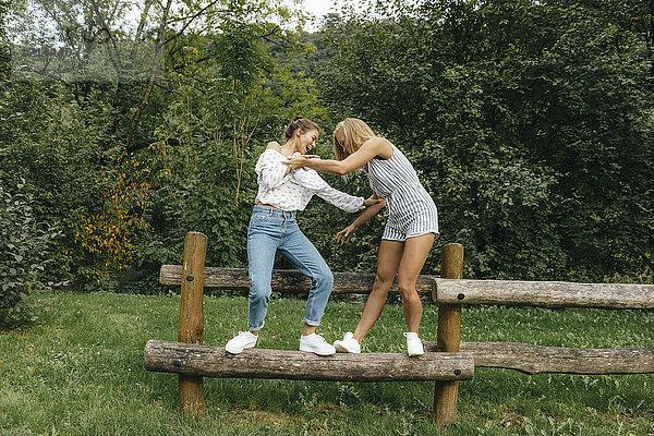 Zwei glückliche junge Frauen  die auf einem Zaun in einem Park balancieren.