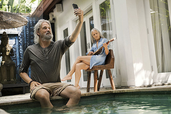 Senior Mann nimmt Selfie am Pool  während seine Frau Gitarre spielt.