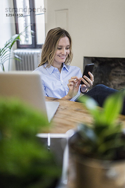 Lächelnde Frau sitzt zu Hause am Schreibtisch und überprüft das Handy