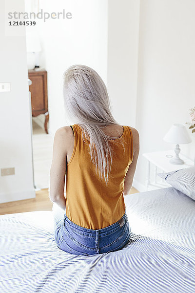 Rückansicht einer jungen Frau mit langen blonden Haaren  die zu Hause auf dem Bett sitzt.