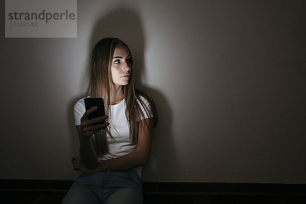 Junge Frau zu Hause auf dem Boden sitzend mit dem Handy im Dunkeln