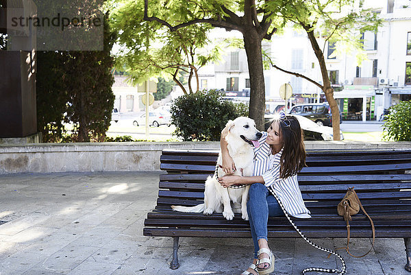 Glückliche junge Frau sitzt mit ihrem Hund auf der Bank in der Stadt.