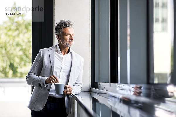 Lächelnder reifer Geschäftsmann mit Kaffeetasse aus dem Fenster schauend