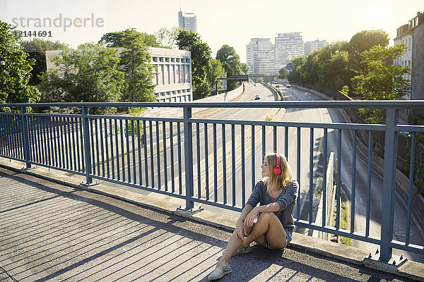 Junge Frau sitzt auf der Brücke und hört Musik mit Kopfhörern.