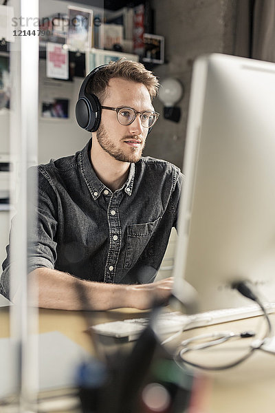 Junger Mann mit Kopfhörern arbeitet am Computer zu Hause am Schreibtisch