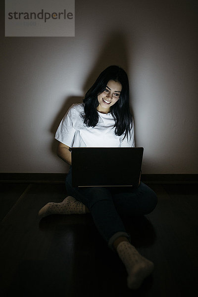 Junge Frau zu Hause auf dem Boden sitzend mit Laptop im Dunkeln