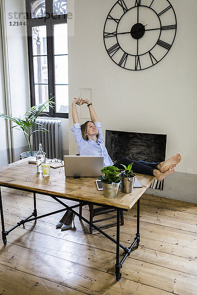 Entspannte Frau sitzt zu Hause am Schreibtisch mit erhobenen Füßen