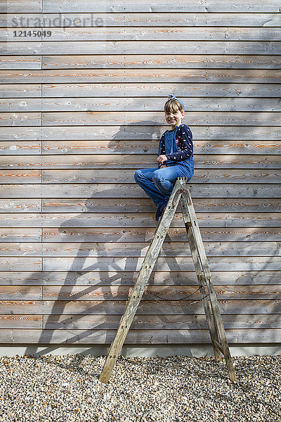 Lachendes Mädchen auf einer Leiter vor einer Holzfassade sitzend