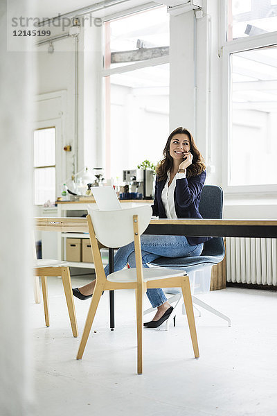 Porträt einer lachenden Geschäftsfrau am Telefon  die am Schreibtisch in einem Loft sitzt.