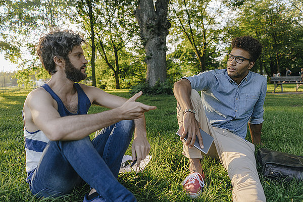 Zwei Freunde sitzen in einem Park mit mobilem Gerät und Diskussionsbeiträgen