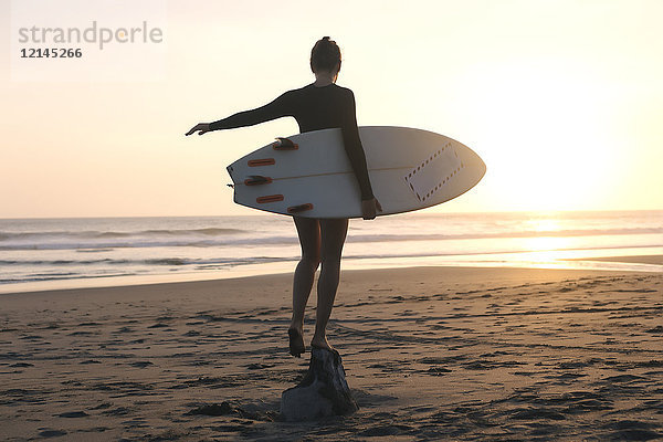Indonesien  Bali  junge Frau mit Surfbrett stehend auf Stein bei Sonnenuntergang