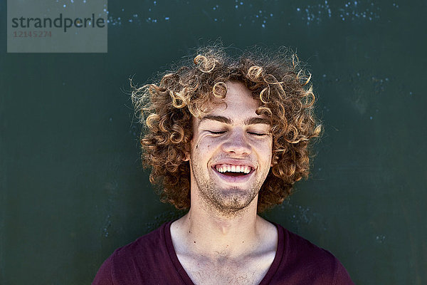 Porträt eines lachenden jungen Mannes mit lockigen Haaren vor einer grünen Wand