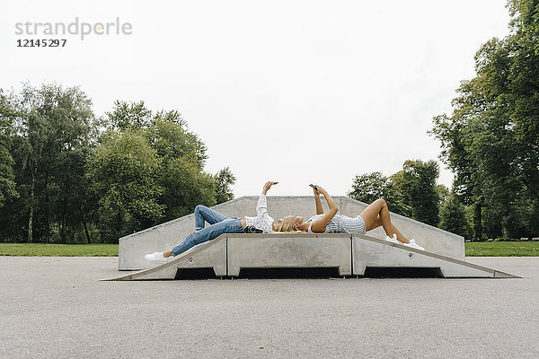 Zwei junge Frauen mit Handys liegen auf einer Rampe in einem Skatepark.