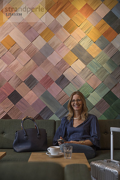 Porträt einer lachenden Geschäftsfrau mit Gepäck und Handy im Cafe