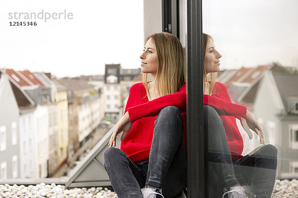 Lächelnde junge Frau sitzt am Fenster in der Stadtwohnung