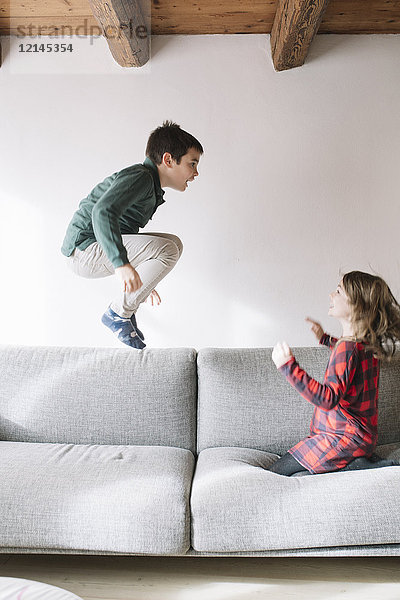Junge  der auf der Couch in die Luft springt  während seine Schwester ihn beobachtet.