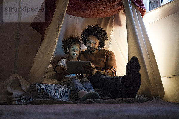 Vater und Tochter sitzen im dunklen Kinderzimmer und sehen sich das digitale Tablett an.