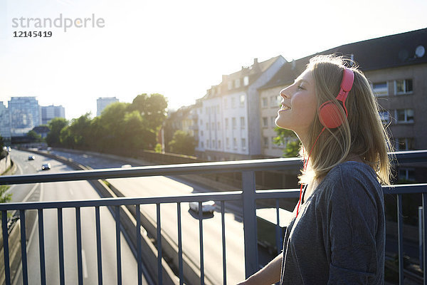 Lächelnde junge Frau auf einer Brücke beim Musikhören mit Kopfhörern