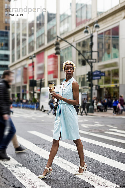 USA  New York  junge blonde Afro-Amerikanerin mit Tasse Kaffee und Smartphone überquert Straße