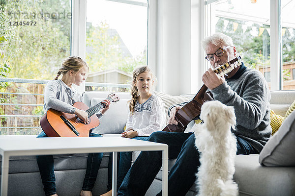 Zwei Mädchen und Großvater auf dem Sofa beim Gitarrespielen mit Hundebeobachtung