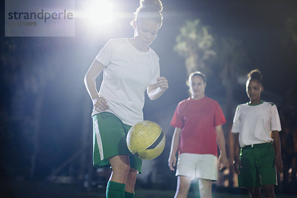Fokussierte junge Fußballspielerin  die nachts auf dem Spielfeld trainiert und den Ball in die Knie zwingt.