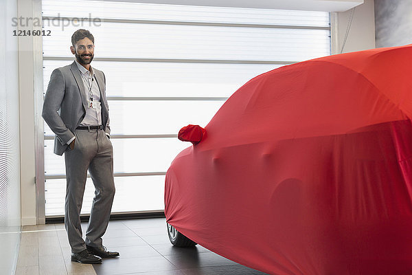 Portrait selbstbewusster Autoverkäufer neben dem gedeckten Auto im Autohaus-Showroom