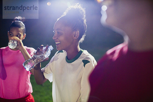 Lächelnde junge Fußballspielerinnen ruhen sich aus und trinken nachts aus Wasserflaschen auf dem Spielfeld.