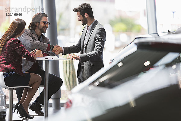 Autoverkäufer beim Händeschütteln mit zwei Kunden am Tisch im Autohaus-Showroom