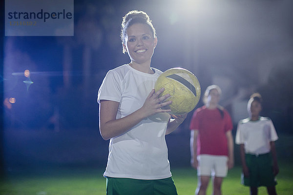 Porträt einer lächelnden  selbstbewussten jungen Fußballspielerin  die nachts den Ball auf dem Spielfeld hält.