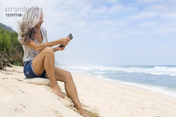Schöne lächelnde Seniorin sitzt am Strand  hält E-Book