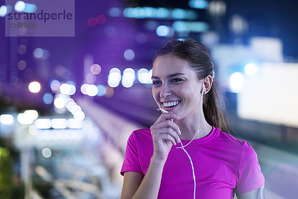 Lächelnde junge Frau im rosa Sportshirt beim Musikhören in der Stadt bei Nacht