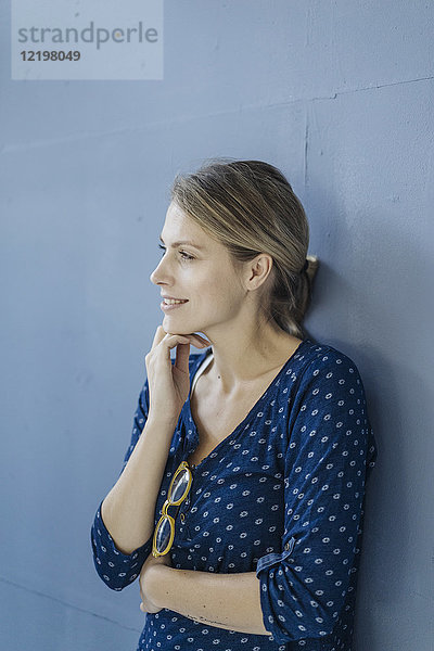 Porträt einer lächelnden Frau  die sich an die blaue Wand lehnt und in die Ferne schaut.