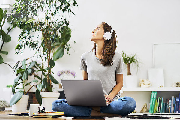 Entspannte junge Frau zu Hause  die mit dem Laptop auf dem Boden sitzt und Musik hört.