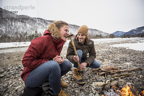Glückliches Paar auf einer Reise im Winter beim Würstchenessen am Lagerfeuer