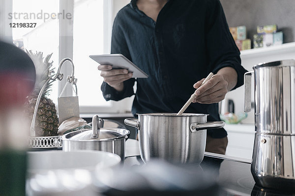 Mann steht in der Küche und liest Nachrichten auf seinem digitalen Tablett  während er kocht.