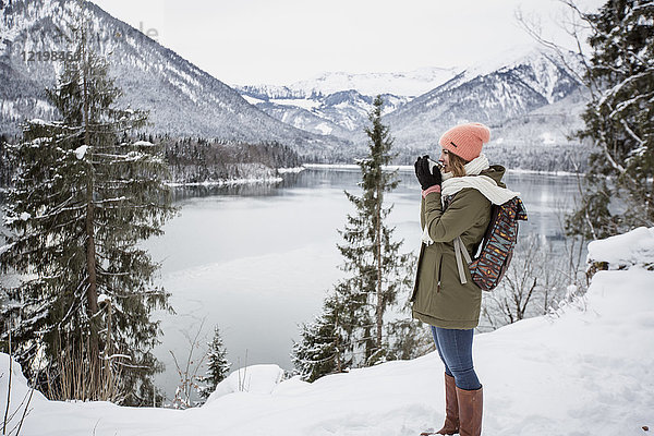 Junge Frau mit heißem Getränk stehend in alpiner Winterlandschaft mit See