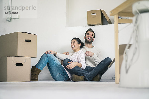 Lachendes Paar sitzt in neuem Zuhause  umgeben von Pappkartons