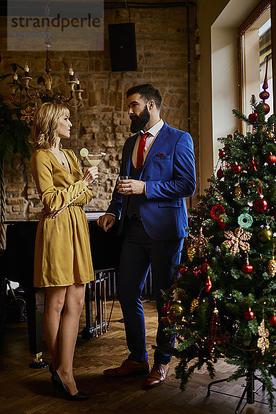 Elegantes Paar mit Getränken am Weihnachtsbaum stehend