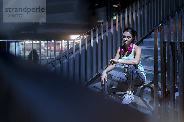 Junge Frau mit rosa Kopfhörern sitzt auf der Treppe und überprüft ihre Smartwatch