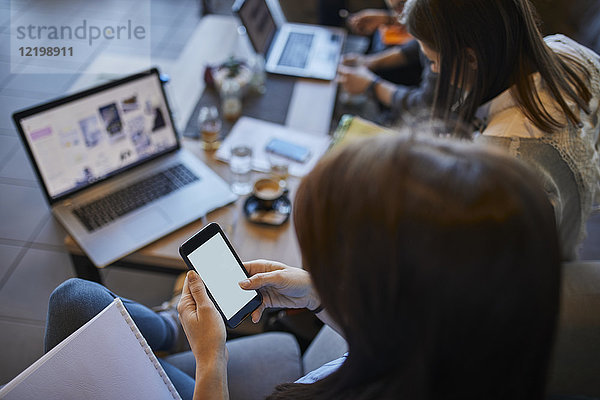 Junge Frau im Café mit Smartphone und Freunden mit Laptop im Hintergrund
