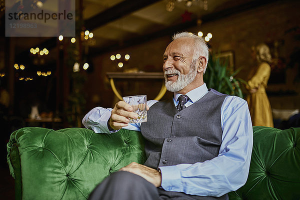 Porträt eines eleganten älteren Mannes  der auf einer Couch in einer Bar mit Becher sitzt.