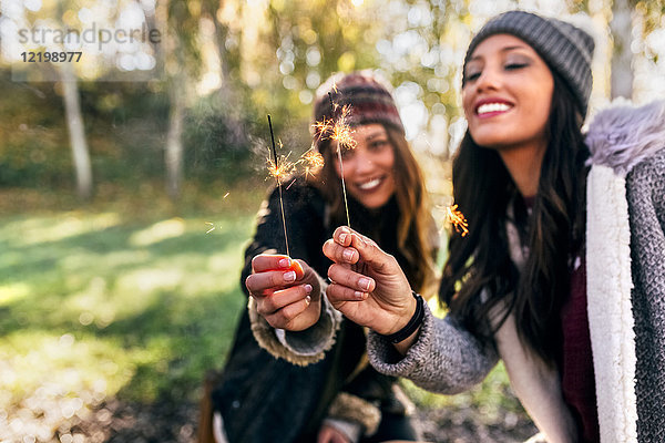 Zwei fröhliche Frauen halten Wunderkerzen in einem herbstlichen Wald.