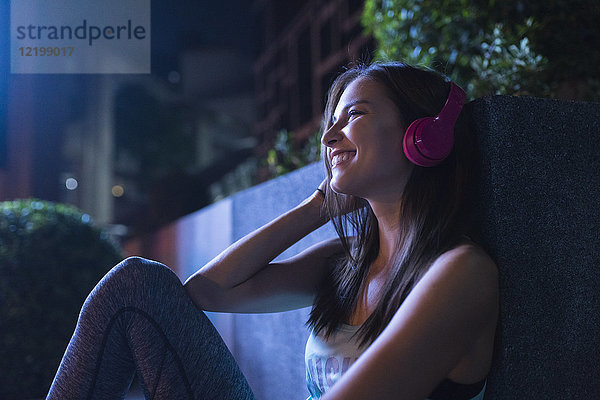 Fröhliche junge Frau mit rosa Kopfhörern  die nachts in der modernen Stadt Musik hört.