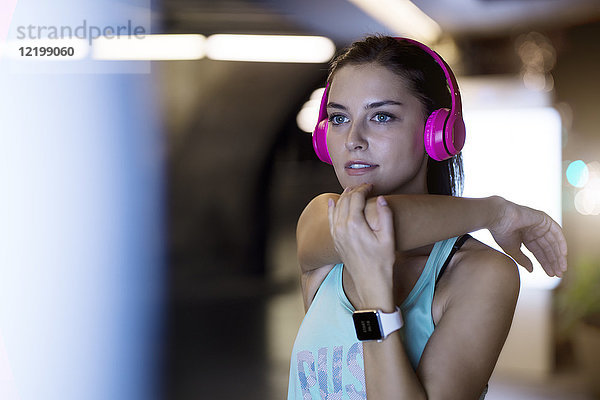 Junge Frau mit rosa Kopfhörern beim Stretching und Musikhören in moderner urbaner Umgebung bei Nacht