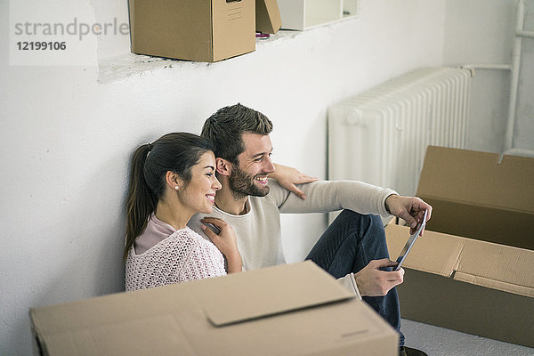 Ein Paar sitzt in einem neuen Zuhause  umgeben von Pappkartons und schaut auf das Tablett.