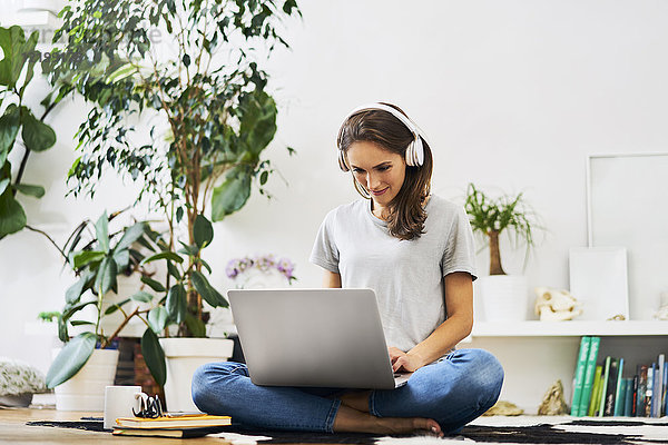 Junge Frau zu Hause sitzend auf dem Boden mit Laptop und Musik hören